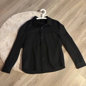 Svart overshirt från Zara i tjockt stretchigt material  - Stilren svart overshirt från Zara med klassisk krage, två bröstfickor och knäppning framtill. Skjortan har lång ärm och rak passform, perfekt att bära öppen eller stängd. Materialet känns mjukt och har en lätt struktur.