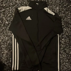 Svart Adidas ziptröja med vita ränder - Svart Adidas ziptröja med klassiska vita ränder längs ärmarna och vit logga på bröstet. Hög krage, hel dragkedja och vita detaljer på axlarna. Perfekt för sport eller chill. Tillverkad i mjukt och stretchigt polyestermaterial.
