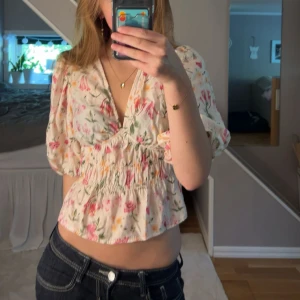 Blommig blus med puffärm och v-ringning - Supersöt blommig blus i ljusa färger med rosa och gröna detaljer. Blusen har v-ringning, puffiga korta ärmar och smockad midja som ger en fin form. Perfekt för vår och sommar, till jeans eller kjol. Materialet känns lätt och luftigt.
