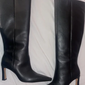 Svarta höga stövlar med klack - Snygga svarta stövlar från NA-KD med spetsig tå och hög klack. Tillverkade i slätt skinnmaterial och har en diskret dragkedja på insidan för enkel påtagning. Stövlarna är knähöga och har en stilren, elegant look som passar till många outfits. Kommer med original paketering. Sälja för att det inte används mer har använt dem två gånger. 