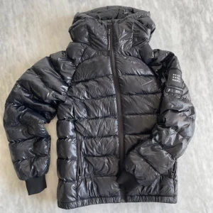 Glansig peak jacka - Snygg svart dunjacka från Peak Performance med glansig finish och huva. Jackan har dragkedja framtill, ribbade muddar och diskret logga på ärmen. Perfekt för kalla dagar och riktigt skön att bära.