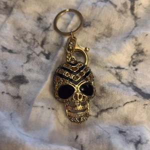 Guldigt dödskalle-hänge med svarta detaljer - Unikt dödskalle-hänge i guld med svarta stenar och glittriga detaljer. Perfekt som smycke eller nyckelring för dig som gillar edgy stil. Hängs enkelt på väskan eller nyckelknippan med den medföljande ringen och klämman.