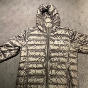 Grå pufferjacka från EA7 Emporio Armani - Säljer en grå pufferjacka från EA7 Emporio Armani med huva och dragkedja framtill. Jackan har ett stilrent quiltat mönster och logga på bröstet. Perfekt för kalla dagar när du vill hålla stilen och värmen.