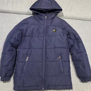 Mörkblå pufferjacka från Lyle & Scott - Mörkblå pufferjacka från Lyle & Scott med huva och dragkedja framtill. Jackan har två fickor med dragkedja och den klassiska gula loggan på bröstet. Perfekt för kyliga dagar och riktigt snygg streetstil.