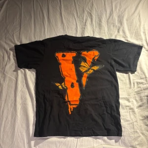Juice wrld Vlone t-shirt - Svart t-shirt från VLONE med trycket 'Legends Never Die' på bröstet och stor orange V med fjärilar på ryggen. Klassisk rund hals och korta ärmar. Skön streetwear-vibe och tillverkad i mjuk bomull.  