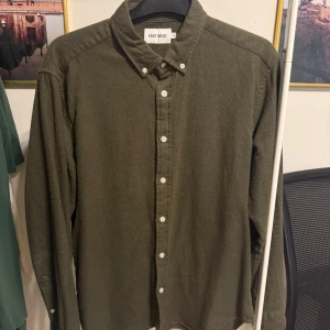 Olivgrön skjorta från East West - Olivgrön skjorta från East West med klassisk krage och vita knappar hela vägen. Skjortan är långärmad och har en avslappnad passform. Tillverkad i mjuk bomull som känns skön mot huden. Perfekt för en stilren och enkel look.