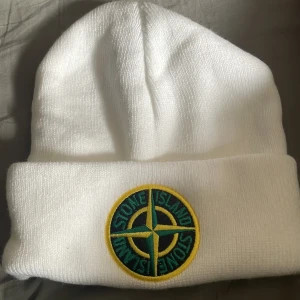 Vit Stone Island mössa med patch - Vit ribbstickad mössa från Stone Island med klassisk rund patch framtill i grönt, gult och svart. Mössan har uppvikt kant och är tillverkad i mjukt stickat material som håller dig varm och snygg under kalla dagar.