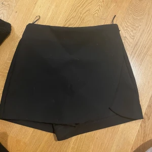 Svart omlott kortkjol med shorts - Svart kortkjol med omlott-design och inbyggda shorts. Kjolen har en stilren look med raka linjer och två fuskfickor bak. Perfekt för dig som vill ha kjolens stil men shortsens komfort. Enkel att matcha med allt.