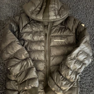 Svart Peak Performance dunjacka - Säljer en svart dunjacka från Peak Performance med huva och dragkedja framtill. Jackan har quiltad design och är perfekt för kyliga dagar. Loggan sitter på bröstet och axeln. Jackan är lätt och har en snygg, sportig look.