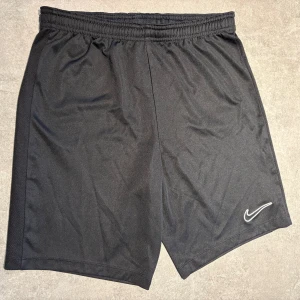 Svarta Nike Dri-FIT shorts - Svarta sportiga shorts från Nike med Dri-FIT-teknologi. De har elastisk midja och en klassisk Nike Swoosh-logga på benet. Perfekta för träning eller chill dagar. Lätta och svala i syntetmaterial.