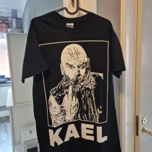 Svart KAEL t-shirt med tryck och autograf - Svart t-shirt med stort vitt tryck av en skäggig man som visar fingret och texten KAEL under. Trycket är signerat med en autograf i guld ovanpå motivet. Klassisk passform och rund hals. Perfekt för dig som gillar unika och edgy plagg.