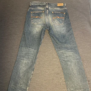 Nudie Jeans Sleepy Sixten blå 33/32 - Blå jeans från Nudie Jeans, modell Sleepy Sixten Celestial Orange. Klassisk femficksdesign med raka ben och orangea sömmar. Tillverkade i 100% bomull för en autentisk jeanskänsla. Perfekt för dig som gillar en avslappnad och tidlös look. Kanske dem mest trendiga jeansen på marknaden just nu!