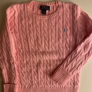 Rosa kabelstickad tröja Polo Ralph Lauren - Supersöt ljusrosa kabelstickad tröja från Polo Ralph Lauren med rund halsringning och klassisk broderad logga på bröstet. Tröjan har långa ärmar och ribbade muddar för en snygg passform. Perfekt för lager-på-lager under hösten och vintern. BARN STORLEK 18-20 ÅR