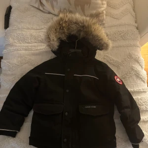  Canada Goose Barn Ny! - Svart dunjacka från Canada Goose med fluffig päls på huvan och klassisk logga på ärmen. Jackan har stora fickor framtill, vita detaljer och knappar längs öppningen. Använd max 10 gånger köpt från Canada goose hemsida för 5år sen för 7995kr, samma jacka idag kostar 12.000kr.. säljes nu för endast 2500kr. Ålder 4-5 i jackan men passar 3-6år. Unik jacka o svår att få tag på! 