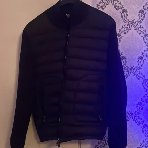 Svart cardigan jacka från Moncler M - Säljer en svart cardigan från Moncler i storlek M. Jackan har quiltad front, släta ärmar och dragkedja framtill. Klassisk Moncler-logga på ena ärmen. Perfekt för dig som vill ha en stilren och snygg jacka med sportig känsla.