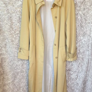 Vintage gul trenchcoat från Kappahl No1 - Snygg gul vintage trenchcoat från Kappahl No1 Collection med klassisk krage och knappar framtill. Jackan har långa ärmar med knappdetaljer vid mudden och en stilren, rak passform. Från 80-talet så ca 40 år. Väldigt bra skick med tanke på åldern, inga defekter.