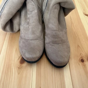 Beige mockastövlar med klack - Snygga beige stövlar i mocka med rund tå och bred klack. Stövlarna har dragkedja på insidan och en mjuk, följsam känsla. Perfekta för dig som vill ha en stilren look med lite höjd. Passar till både jeans och kjol.