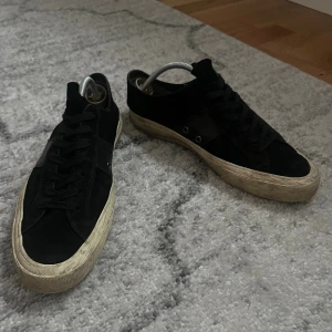 Svarta Tom Ford sneakers i mocka - Svarta sneakers från Tom Ford i mjuk mocka med klassisk låg profil och rund tå. Skorna har snörning och en beige, något smutsad gummisula med Tom Ford-logga baktill och under sulan. Perfekta för dig som gillar stilrena och exklusiva sneakers.
