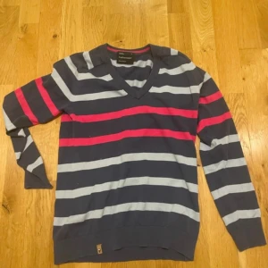 Randig Peak Performance tröja, navy - Randig tröja från Peak Performance i marinblått med ljusgrå och rosa ränder. V-ringad modell med långa ärmar och ribbade muddar. Perfekt för dig som gillar klassisk stil med en färgklick. Materialet är mjuk bomull.