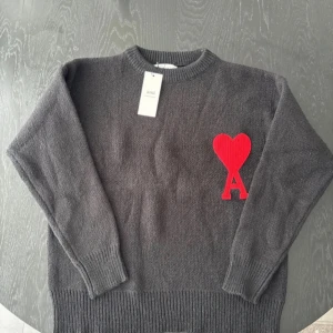Svart stickad tröja från Ami - Mysig grå stickad tröja från Ami med rund hals och ribbade muddar. På bröstet sitter en stor röd hjärtapplikation med bokstaven A. Perfekt för dig som vill ha en stilren men ändå unik look.