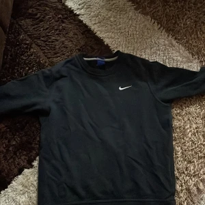 Svart Nike sweatshirt med logga - Svart sweatshirt från Nike i storlek M med klassisk vit swoosh-logga på bröstet. Tröjan har rund hals och långa ärmar, perfekt för chill dagar eller när du vill ha en enkel och stilren look. Materialet är mjukt och känns skönt mot huden.