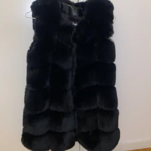 Svart fluffig pälsväst - Mysig svart pälsväst med breda, markerade paneler och hög krage. Västen är lång och har en riktigt fluffig look som ger en lyxig känsla. Perfekt för att addera extra edge till din outfit under kyliga dagar.