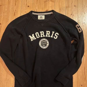 Marinblå stickad tröja från Morris Gents - Marinblå stickad tröja från Morris Gents med vita bokstäver och tryck framtill. Snygga grå armbågslappar och siffran 68 på ena ärmen. Klassisk rund halsringning och ribbade muddar. Perfekt för dig som gillar sportig och stilren look.