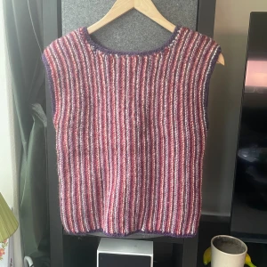 Randig stickad väst i lila och rött - Säljer en mysig stickad väst med vertikala ränder i lila, rött och vitt. Västen har en rund halsringning med lila kant och är ärmlös. Står ingen storlek angivet med uppskattr S/M