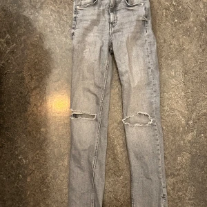 Grå ripped jeans från Zara - Säljer ett par grå jeans från Zara i storlek 34. Byxorna har raka ben, slitna hål vid knäna och klassisk femficksdesign. Storlek 34
