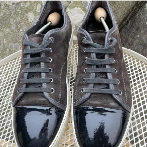 Lanvin sneakers i mocka och lack - Snygga sneakers från Lanvin i mörkgrå mocka med svart lackad tå och vita sulor. Skorna har klassisk snörning och rund tå, vilket ger en clean och lyxig vibe. Perfekta för dig som gillar stilrena detaljer och exklusiva material.