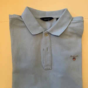 Ljusblå pikétröja från GANT - Snygg ljusblå pikétröja från GANT med klassisk krage och två knappar framtill. Tröjan har korta ärmar och GANT:s broderade logga på bröstet. Tillverkad i bomull för en skön och luftig känsla. Perfekt för en clean och stilren look. 