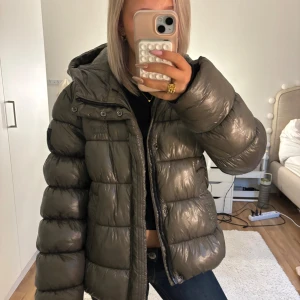 Grå pufferjacka från Superdry XP-D - Säljer en grå pufferjacka från Superdry XP-D med glansig finish och stor huva. Jackan har quiltad design, dragkedja framtill och svart patch på ärmen. Perfekt för kalla dagar och riktigt snygg till streetwear-stilen. Säljer den för den är förstor för mig i armarna då den är XL men tycker den passar bra ändå annars för någon som har XS och är 156 cm, köpte för 2000kr