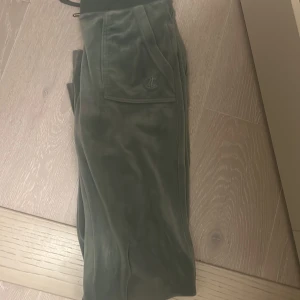 Gröna mjukisbyxor Juicy Couture XS - Mysiga gröna mjukisbyxor från Juicy Couture i velour med snörning i midjan och broderad logga på fickan. Byxorna har bred resår i midjan och klassisk relaxed fit, perfekta för chill dagar.