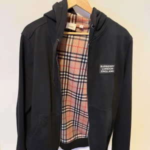 Svart Burberry hoodie med rutig insida - Svart hoodie från Burberry med klassiskt rutigt foder i beige, rött och svart. Tröjan har dragkedja, huva med snörning och Burberry London England-tryck på bröstet. Perfekt för dig som vill ha en stilren men ändå lyxig look.