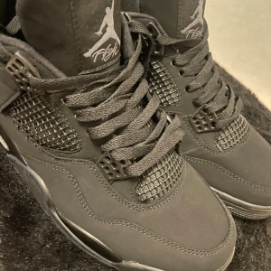 Nike Air Jordan 4 Retro Black cat - Säljer ett par helsvarta Nike Air Jordan 4 Retro sneakers med ikoniska Jumpman-loggor på plös och häl. Skorna har meshpaneler, snörning och en robust sula med synlig Air-enhet. Ovandelen är i mocka och detaljer i syntet, vilket ger en clean och sportig look.