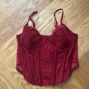 Vinröd spetskorsett från H&M - Säljer en vinröd korsett från H&M med blommig spets och justerbara axelband. Korsetten har markerade sömmar och meshdetaljer som ger en snygg siluett. Perfekt för dig som vill ha en statement topp med romantisk vibe.
