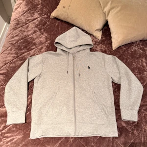 Grå Polo Ralph Luaren Zip Hoodie - Säljer denna stiliga Gråa Ralph Lauren hoodie med dragkedja. Den har aldrig använts då den är för stor för mig och är i suverän skick. Storlek på varan är M. Hör av dig vid intresse! 