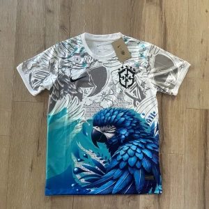 Nike x CBF t-shirt med blå papegoja - Unik t-shirt från Nike x CBF med stor blå papegoja och gråa grafiska detaljer. T-shirten har korta ärmar, rund hals och är tillverkad i lätt polyester. Färgerna går i vitt, blått och grått med coolt tryck och CBF-logga på bröstet.