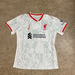 Liverpool FC vit bortatröja Nike - Liverpool FC bortatröja från Nike i vit med ljusgrått mönster. Kortärmad med röd och gul detalj på krage och ärmslut. Broderat klubbmärke och Nike-logga på bröstet, samt sponsortryck. Tillverkad i lätt, ventilerande Dri-FIT-material.