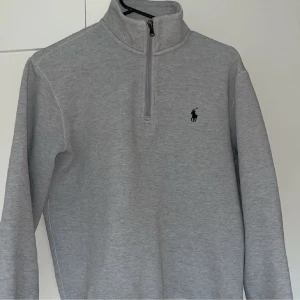 Grå half zip tröja från Polo Ralph Lauren - Snygg ljusgrå långärmad tröja från Polo Ralph Lauren med klassisk svart logga på bröstet. Tröjan har en hög krage och half zip-dragkedja framtill. Perfekt för lager-på-lager och chill stil. Tillverkad i mjuk bomullsblandning.