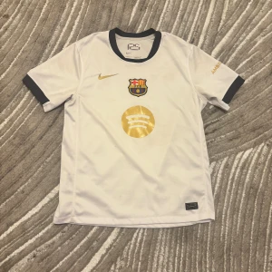 Vit FC Barcelona Nike fotbollströja - Säljer en vit FC Barcelona fotbollströja från Nike med guldiga detaljer och klubbmärke på bröstet. Tröjan har korta ärmar med mörkblå muddar och är tillverkad i lätt, ventilerande Dri-Fit material. Snyggt Spotify-tryck i guld på framsidan och mörkblå krage.