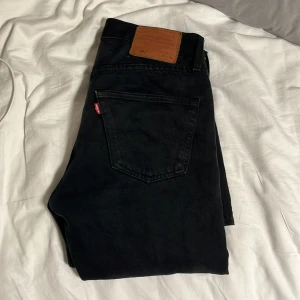 Svarta Levi's 501 jeans W30 L34 - Svarta Levi's 501 jeans med klassisk rak passform och fem fickor. Jeansen har knappgylf, detaljer i kopparfärg och den ikoniska läderpatchen bak i midjan. Materialet är slitstarkt denim och färgen är djup svart, perfekt för en clean och stilren look.