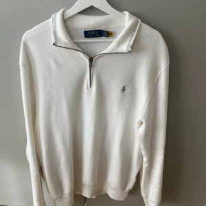 Vit half zip tröja från Polo Ralph Lauren - Vit långärmad tröja från Polo Ralph Lauren med half zip-dragkedja och klassisk krage. Tröjan har diskret broderad logga på bröstet och är tillverkad i bomull med en stickad känsla. Perfekt för en clean och stilren look.
