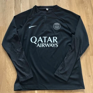 Svart PSG fotbollströja Nike S - Säljer en svart långärmad fotbollströja från Nike med PSG-logga och Qatar Airways-tryck på bröstet. Tröjan har halv dragkedja fram och är tillverkad i ett lätt, funktionellt material som andas. Perfekt för träning eller match.