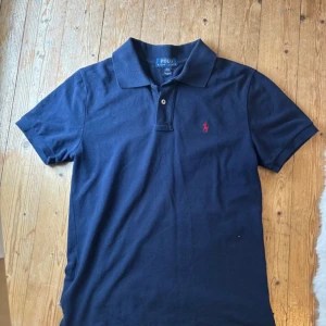 Marinblå pikétröja från Ralph Lauren - Snygg marinblå pikétröja från Polo Ralph Lauren i storlek L i barn 158 cm. Klassisk krage, korta ärmar och två knappar framtill. Tröjan har det ikoniska röda Polo-logot broderat på bröstet. Tillverkad i mjuk bomull som är skön mot huden.