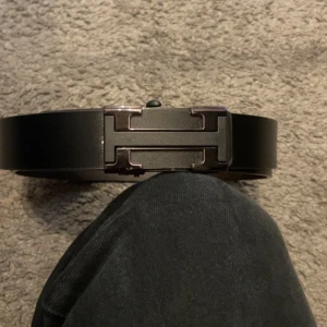 Svart skärp med metallspänne - Snyggt svart bälte med ett stilrent, rektangulärt metallspänne i mörk finish. Bältet har en slät yta och är tillverkat i skinn, vilket ger en klassisk och clean look. Perfekt för att lyfta din outfit med en modern touch.