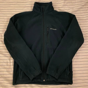 Mörkblå fleecejacka från Columbia - Mörkblå fleecejacka från Columbia med hög krage och hel dragkedja framtill. Jackan har en bröstficka med dragkedja och två sidofickor. Perfekt för kyliga dagar och outdoor-aktiviteter. Stilren och enkel design med Columbia-logga på bröstet.