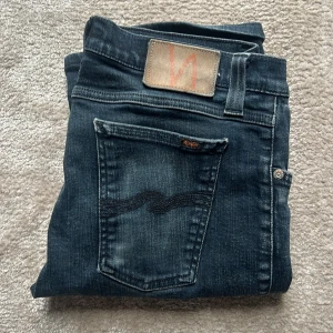 Nudie jeans - Säljer ett par mörkblå jeans från Nudie Jeans med klassisk design och snygga detaljer på bakfickorna. Jeansen har en smal passform och är tillverkade i mjukt denimtyg med lätt tvättad utseende.