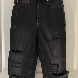 Svarta ripped jeans från H&M Divided - Svarta jeans från H&M Divided i storlek 36 med flera slitningar och hål på benen för en edgy look. Jeansen har rak passform, hög midja och råa benslut. Tillverkade i 100% bomull för en klassisk jeanskänsla.