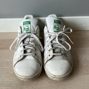 Adidas Stan Smith vita sneakers - Klassiska Adidas Stan Smith sneakers i vitt med gröna detaljer på häl och plös. Skorna har snörning, perforerade sidor och platt sula. Tillverkade i skinn. Perfekt för dig som gillar retrostil och ikoniska modeller. Passar både killar och tjejer.   Pris kan diskuteras